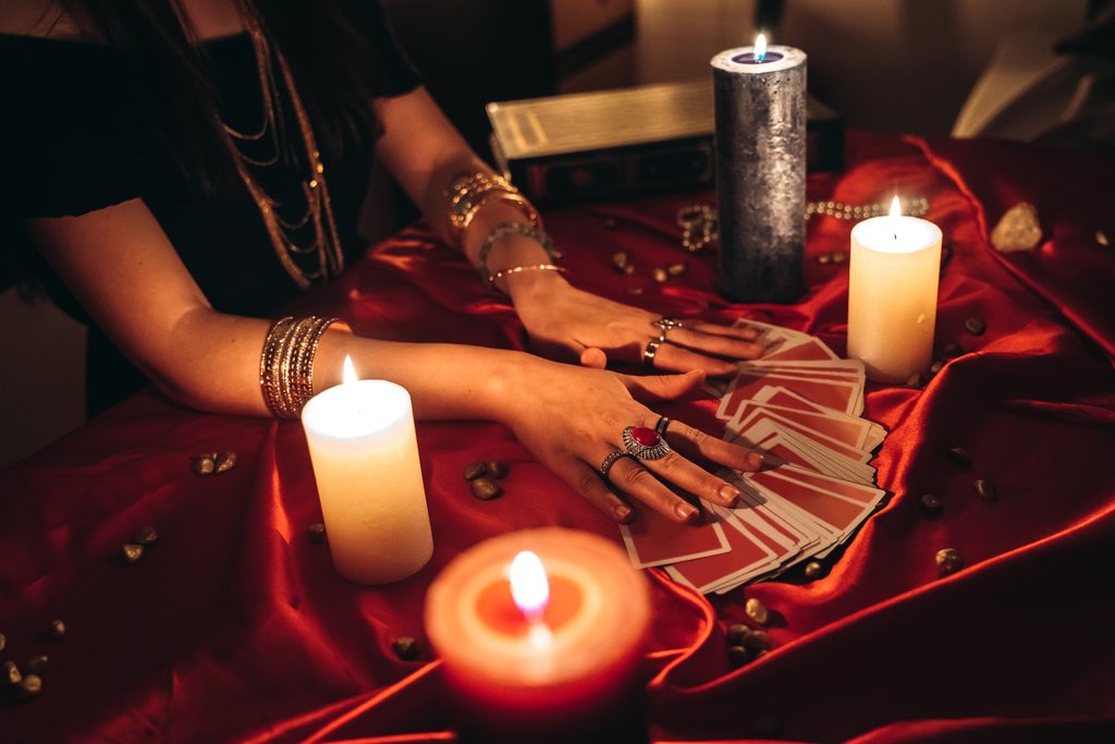 A fortune-teller extends her hands over an array of cards on a red tablecloth, surrounded by candles